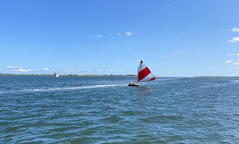 sailboat with a red and white sail in Barnstable Harbor with the Sandy Neck Lighthouse in the background